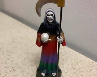 May include: A figurine of the Grim Reaper, holding a scythe and a white orb. The figure wears a black hood and a colorful robe that transitions from red to purple to green. The scythe is gold-colored.