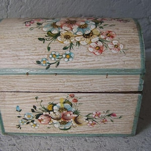 May include: A wooden box with a hinged lid, painted with a floral design in shades of pink, white, and green. The box has a light wood finish and a blue trim.