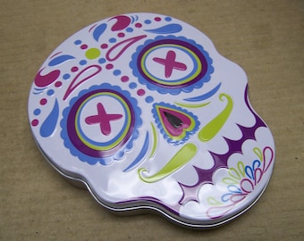May include: A white metal skull-shaped tin with a colorful Day of the Dead design. The skull has purple, blue, yellow, and pink details.