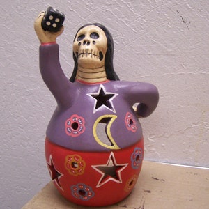 Peut inclure: Figurines en céramique de couleur violette et rouge représentant un squelette tenant un dé noir. La figurine est décorée d'une étoile, d'une lune et de fleurs.