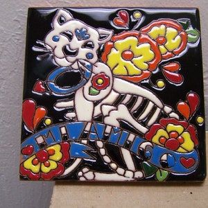 May include: A white ceramic tile with a black background features a colorful Day of the Dead-inspired cat with a skeleton body. The cat is surrounded by flowers and the words "Mi Amigo" in blue and white.