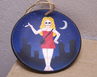 May include: A round ceramic plate with a blue background and white stars. A painted image of a woman with a skull face, blonde hair, and a red dress is on the plate. The woman is holding a cigarette in her right hand and a small black purse in her left hand. A crescent moon is painted in the upper right corner of the plate.