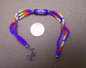 May include: A multi-strand beaded bracelet with purple, orange, yellow, and white beads. The bracelet has a purple and white bead with a geometric design in the center.