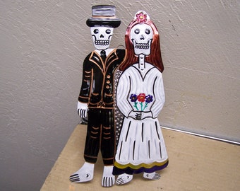 May include: A decorative metal Day of the Dead couple. The male figure wears a black suit and top hat, while the female figure is in a white dress and holds a bouquet of flowers. The figures have skull faces.