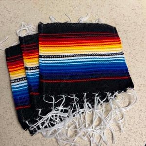 May include: Three rectangular woven fabric mats featuring a black base with horizontal stripes in red, orange, yellow, white, and various shades of blue. Each mat has a fringe of white threads along the bottom edge, creating a textured appearance.