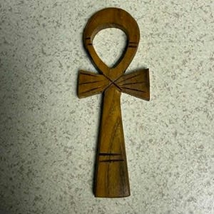 May include: A wooden ankh, a symbol of life, is displayed. The ankh is carved from a light brown wood and features a loop at the top, a horizontal bar, and a vertical stem. The ankh has etched lines for detail.