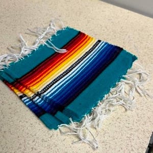 May include: A square woven textile coaster featuring a vibrant striped pattern in shades of teal, red, orange, yellow, white, and dark blue. The fabric has a textured weave and is finished with long, loose white fringe along two opposite edges.