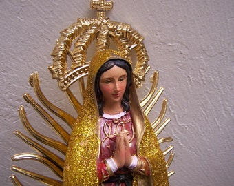 Puede incluir: Una figurita dorada de la Virgen María con una corona y un halo dorado. Lleva una túnica azul con purpurina dorada y un vestido rojo y dorado. Tiene las manos juntas en oración.