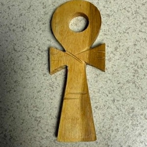 Può includere: Un simbolo ankh in legno, una forma a croce con un anello in cima. L'ankh è scolpito in legno chiaro, che mostra la venatura naturale. Il design presenta una barra orizzontale e una linea verticale che si estende verso il basso.