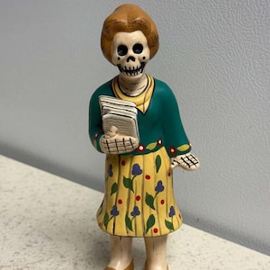 May include: A standing figurine of a skeleton with brown hair, wearing a teal long-sleeved top with a yellow collar and a yellow skirt featuring a blue and red floral pattern. The figure holds a stack of white papers in its right hand.