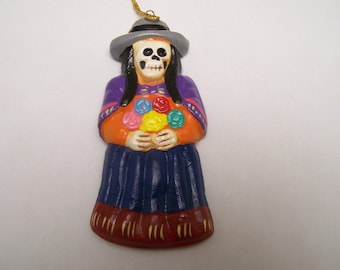 May include: A ceramic ornament depicting a skeleton figure wearing a purple and orange dress, a gray hat, and holding a bouquet of colorful flowers. The ornament is a traditional Day of the Dead decoration.