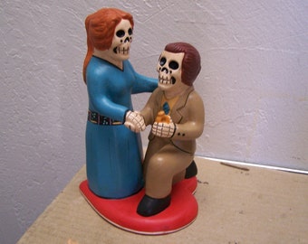 May include: A ceramic figurine of a couple dressed in traditional clothing. The woman is wearing a blue dress and the man is wearing a brown suit. They are both holding hands and smiling. The figurine is painted in bright colors and has a heart-shaped base.