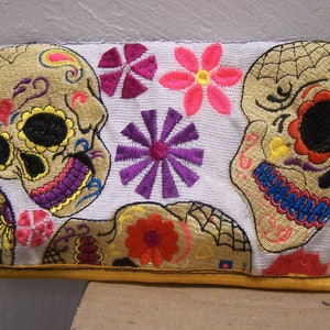May include: White fabric clutch purse with embroidered colorful sugar skulls and floral designs. The purse has a yellow border.