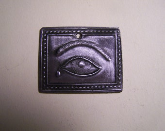 May include: A silver-colored metal pendant with a raised image of an eye with a tear drop. The eye is surrounded by a decorative border.