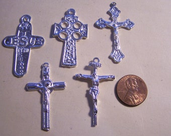 May include: Five silver-toned metal cross charms, including a Celtic cross, a cross with the word "JESUS" on it, and three crucifixes. The charms are arranged on a light brown surface.