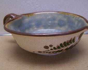 May include: A brown ceramic bowl with a light blue glaze and a green and brown floral design. The bowl has a handle and a spout.