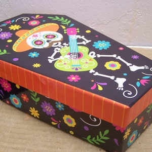 Puede incluir: Una caja negra con forma de ataúd con un diseño colorido del Día de Muertos. La caja presenta un esqueleto tocando una guitarra con un sombrero, flores y espirales.