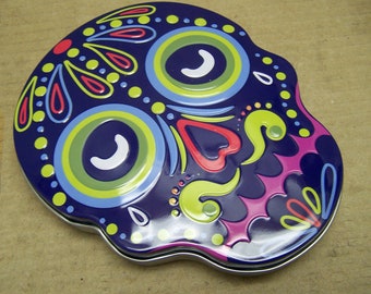May include: A blue metal skull-shaped tin with a colorful Day of the Dead design. The skull has white eyes, a pink nose, and yellow, green, and red details.
