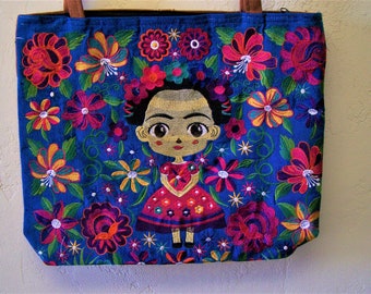 May include: Blue tote bag with an embroidered design featuring a cartoon-style figure surrounded by colorful flowers. The figure has dark hair and is wearing a red dress. The bag has brown handles.