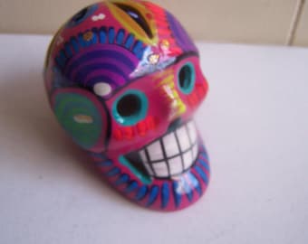 May include: A Day of the Dead-themed ceramic skull figurine. The skull is painted in bright pink, with colorful accents of blue, green, yellow, and purple. The skull has a wide, toothy grin.