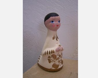 May include: A white ceramic figurine of a person kneeling with their hands clasped in prayer. The figurine is painted with gold details and floral patterns. The person has black hair and blue eyes.