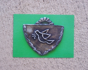 May include: A silver-colored metal ornament shaped like a shield with a dove carrying an olive branch in its beak. The ornament is on a green background.