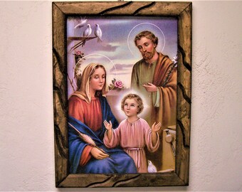 May include: A framed painting depicting the Holy Family. Mary, Joseph, and baby Jesus are depicted in a traditional style with halos around their heads. The painting is set against a background of a garden with a dove perched on a branch.