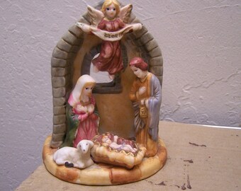 May include: A ceramic nativity scene with a white angel holding a banner that reads "Gloria", a woman in a blue and pink robe, a man in a blue robe, and a baby in a manger with a white lamb.