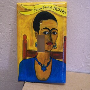 Könnte beinhalten: Ein Lichtschalter-Abdeckung mit einem Gemälde von Frida Kahlo, einer mexikanischen Malerin, mit dem Text "Frida Kahlo 1907-1954". Das Gemälde ist auf einem gelben Hintergrund mit einem gelben Banner.