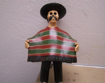 May include: A ceramic figurine of a skeleton wearing a black sombrero and a green and red striped poncho. The figurine has a painted skull face with a mustache.