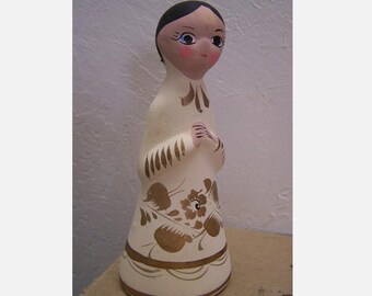 May include: A white ceramic figurine of a woman with black hair, wearing a white dress with gold floral designs. The figurine has painted blue eyes, pink cheeks, and a gold trim around the dress.