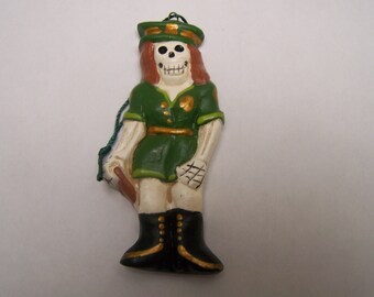 May include: A green and white ceramic ornament of a skeleton wearing a green uniform with gold accents and a black hat. The ornament has a green string attached to the top.