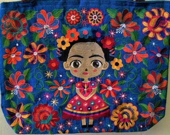 May include: A blue tote bag with a colorful embroidered design featuring a cartoon Frida Kahlo with a flower crown and a red dress. The bag is surrounded by flowers in various colors, including red, orange, yellow, green, and white.