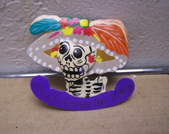 May include: A colorful Day of the Dead figurine of a skeleton wearing a hat with flowers and a purple banner that says "Dia De Los Muertos"
