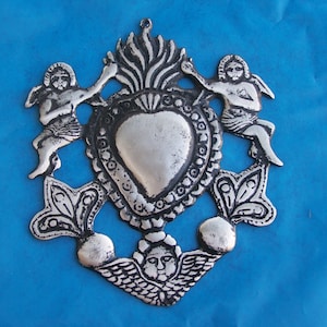 May include: Silver-colored metal ornament depicting a heart with flames and two cherubs. The heart is surrounded by ornate floral designs.