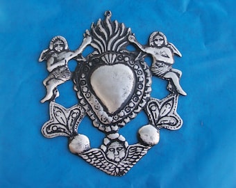 May include: Silver-colored metal ornament depicting a heart with flames and two cherubs. The heart is surrounded by ornate floral designs.