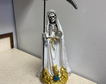 May include: A white statue of a robed skeleton figure holding a scythe and a crystal ball. The figure has a skeletal face and is adorned with gold accents. The base features an owl and a globe.