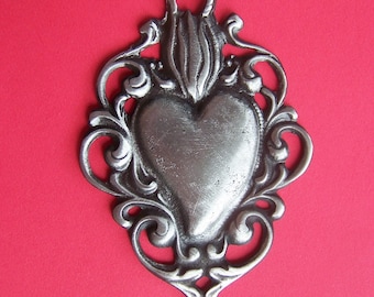 May include: A silver pendant in the shape of a heart with ornate detailing around the edges. The heart is raised and has a smooth surface.
