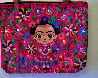 May include: A pink tote bag with a colorful embroidered design featuring a cartoon Frida Kahlo with a floral crown and a pink dress. The bag is surrounded by colorful flowers and leaves.