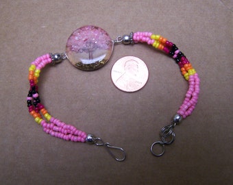 May include: A beaded bracelet with a clear, round charm featuring a pink tree design. The bracelet has three strands of pink beads, accented with yellow, orange, and black beads. The bracelet is secured with a silver clasp.