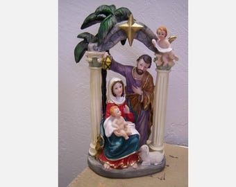 May include: A white ceramic nativity scene with a star above the holy family. The figures are painted in realistic colors and the scene is set under a palm tree archway.