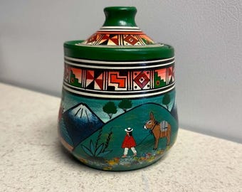 May include: A hand-painted ceramic jar with a lid, featuring a green top with geometric patterns. The body depicts a landscape with a snow-capped mountain, green hills, a person in a red poncho, and a brown donkey, all in vibrant colors.