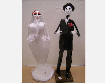 May include: A white wedding dress with a veil and a black tuxedo with a red rose. The figures are made of paper mache and are skeletons.
