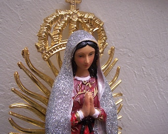 Puede incluir: Una figurita de la Virgen María, vestida con una túnica plateada y blanca con brillantina, y una corona dorada. Ella tiene las manos juntas en oración y tiene una expresión suave en su rostro.