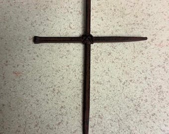May include: A cross formed by two dark brown, textured metal nails joined at the center with a thin, dark cord wrapped around the intersection. The vertical nail features a small circular loop at the top, while the horizontal nail has a flat head.
