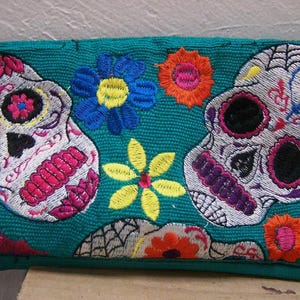 May include: Teal fabric clutch purse with embroidered sugar skulls, flowers, and other designs. The skulls are white with pink, purple, and yellow accents.