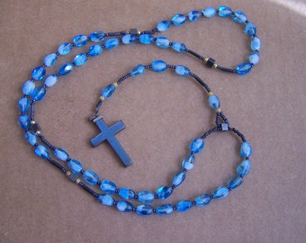 May include: A blue beaded rosary with a silver-colored cross pendant. The beads are oval-shaped and translucent, with a mix of light and dark blue hues. The rosary is laid out on a neutral-toned surface.