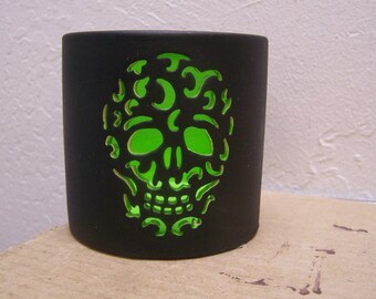 May include: A black cylindrical candle holder with a skull design cut out, revealing a bright green interior. The skull's features are clearly defined, creating a striking contrast. The candle holder is likely made of ceramic or a similar material.