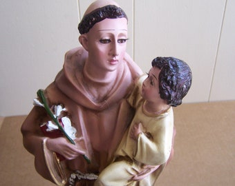 May include: A figurine of Saint Anthony of Padua holding a small child. The saint is wearing a brown robe and a brown cord around his waist. He is holding a bouquet of white flowers in his left hand. The child is wearing a cream-colored robe.