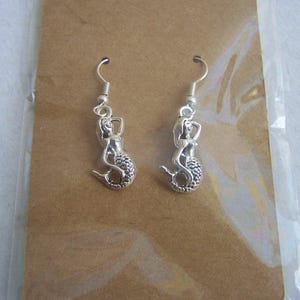 May include: A pair of silver mermaid dangle earrings. The mermaids are in a sitting position with their tails curled around them.